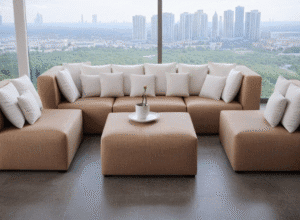 Nowoczesna, beżowa sofa narożna z wieloma białymi poduszkami w salonie z dużymi oknami z widokiem na miasto, oraz kwadratowa beżowa pufa służąca jako stolik - Holiday