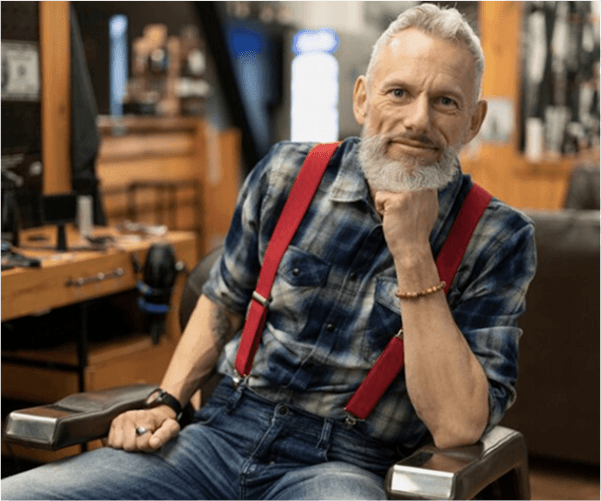 Mężczyzna z siwą brodą, w kraciastej koszuli i dżinsach, siedzący na krześle, z czerwonymi szelkami i bransoletkami na nadgarstkach.