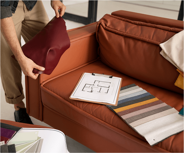 Osoba trzymająca czerwoną, skórzaną poduszkę nad brązową skórzaną kanapą, na której leży clipboard z planem pomieszczenia i próbnik tkanin.