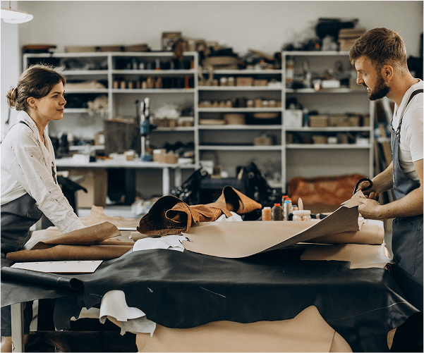 Dwie osoby, kobieta i mężczyzna, pochylone nad dużymi arkuszami materiału w pracowni, z półkami wypełnionymi przedmiotami w tle.