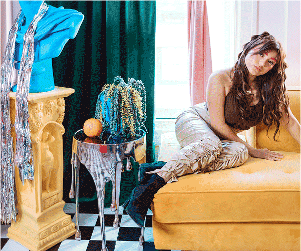 Kobieta z długimi, brązowymi włosami leżąca na żółtej kanapie, w srebrzystych spodniach i brązowym topie, obok stolika z pomarańczą i niebieskim kaktusem, oraz rzeźby na cokole.